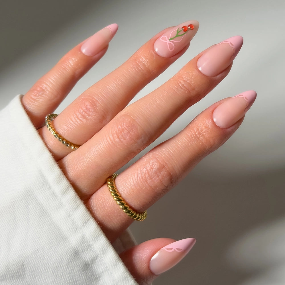 A hand with long, almond-shaped, light pink glamnetic Cherry Bow press-on nails features delicate line art and a cherry accent. Gold rings and a white sleeve complete the chic look of this stylish nail design.