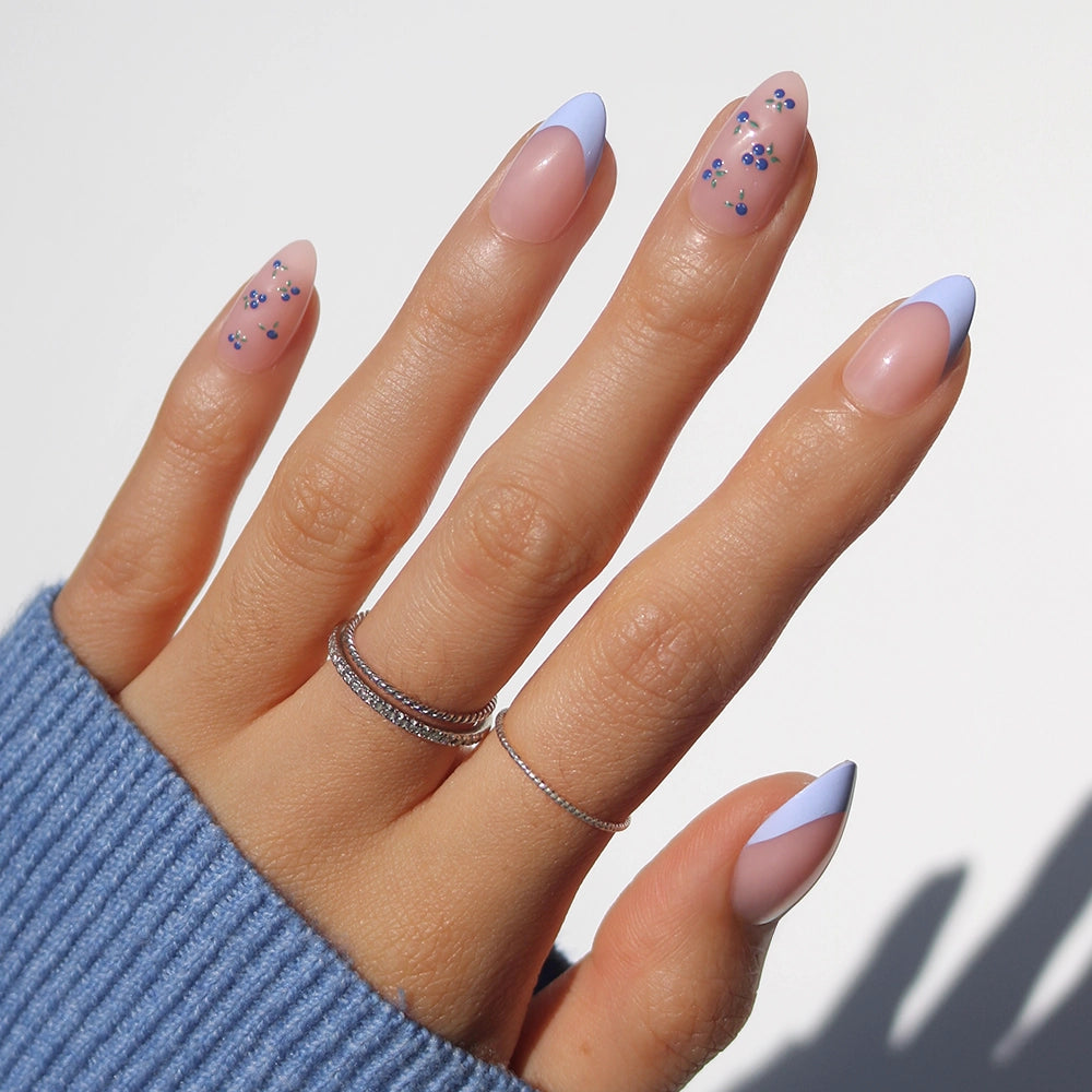 Get inspired by glamnetic’s Bluebelle: almond press-on nails with pastel purple French tips and delicate blue floral accents, shown with silver rings and a blue ribbed sweater—perfect for a chic, stylish look.