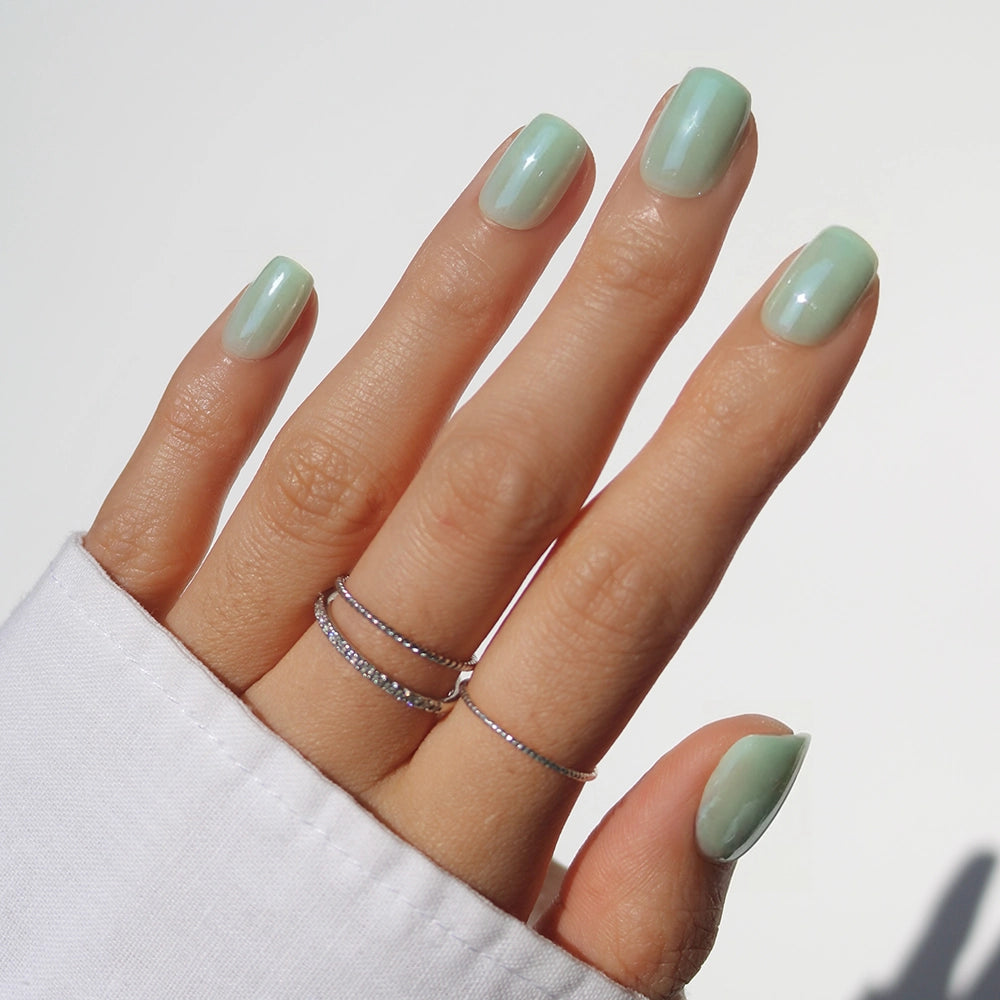 A hand showcases glamnetic’s Honeydewy mint green press-on nails with three delicate silver rings on the middle and ring fingers, set against a white background and paired with a white long-sleeve shirt.