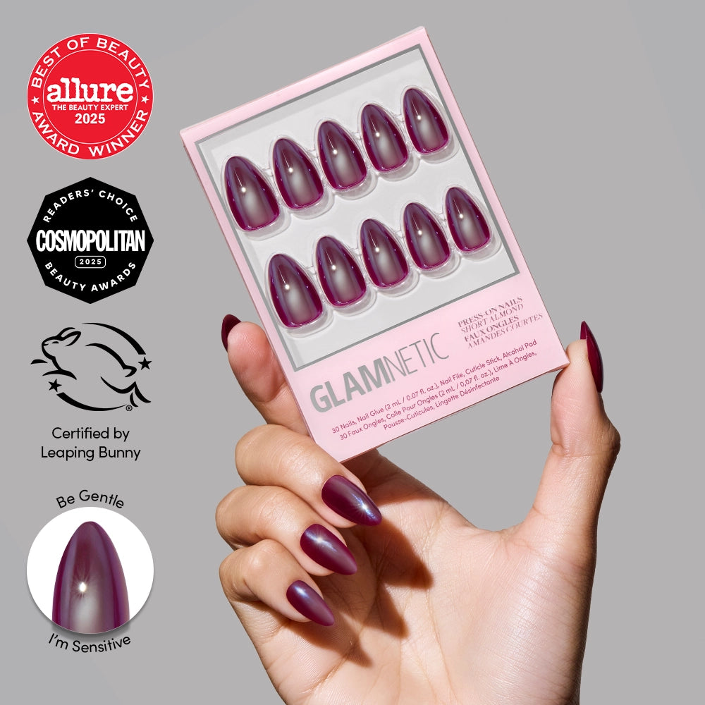 A hand with long, almond-shaped, glossy purple "Jam Drizzle" press-on nails by glamnetic holds a nail kit. Beauty award badges and cruelty-free certification appear on the left. One nail is labeled “Be Gentle, I’m Sensitive.”.