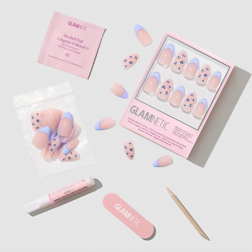 The glamnetic Bluebelle almond nails set shows pastel pink and purple flower designs, pictured with an alcohol pad, nail glue, wooden cuticle stick, nail file, and extra press-ons on a light gray background. - Product image