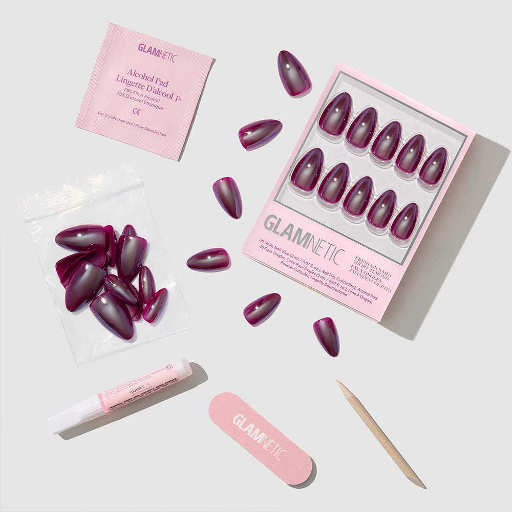 The glamnetic Jam Drizzle set features almond-shaped press-on nails in a pink box, loose glossy purple nails, a pink nail file, wooden cuticle stick, glue, alcohol pad, and resealable bag on a light gray background. - Product image