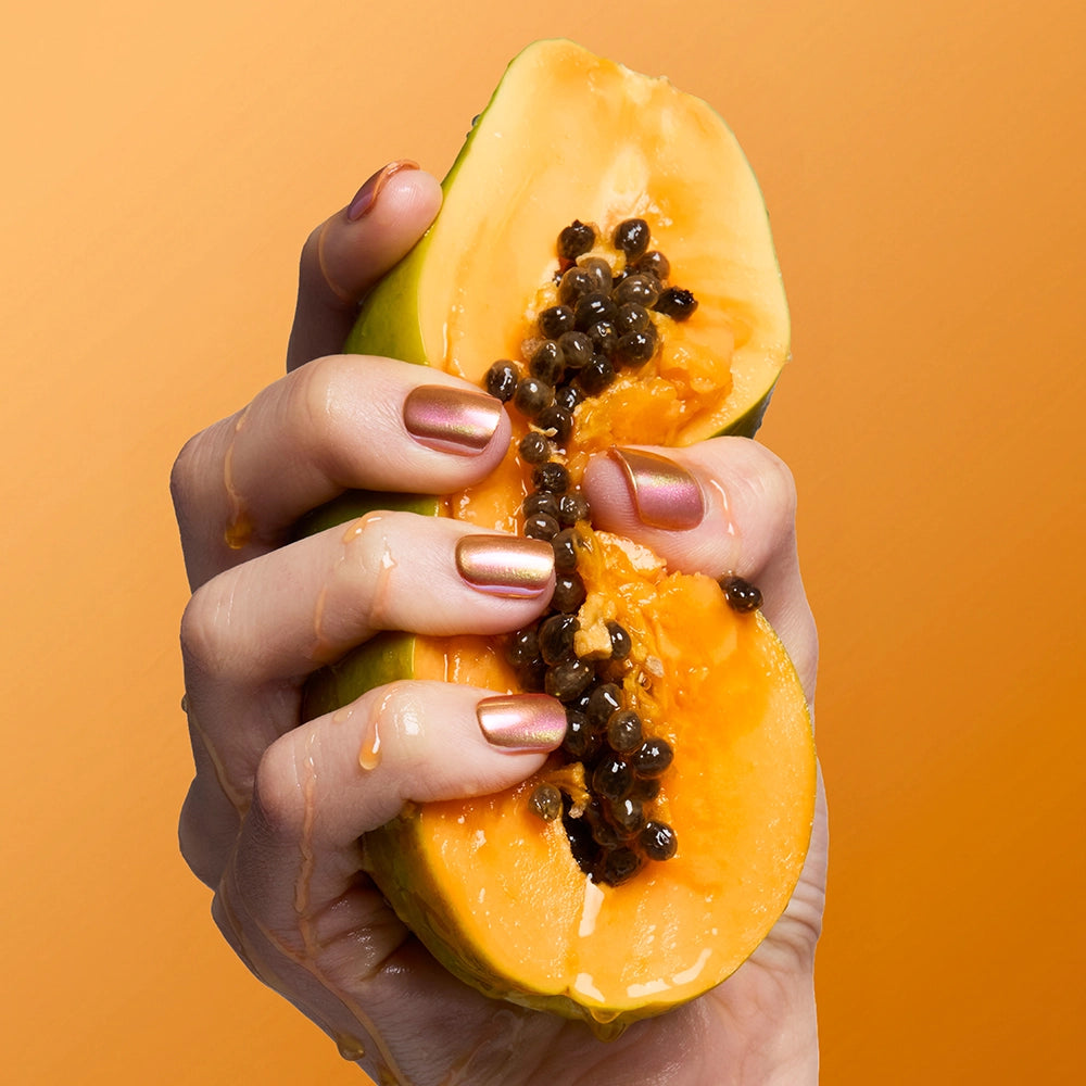 A hand with shiny, orange squoval nails in glamnetic’s Papaya Chrome squeezes a ripe papaya, its juice dripping against a solid orange background and exposing the fruit's vibrant flesh and glossy black seeds. - Product image