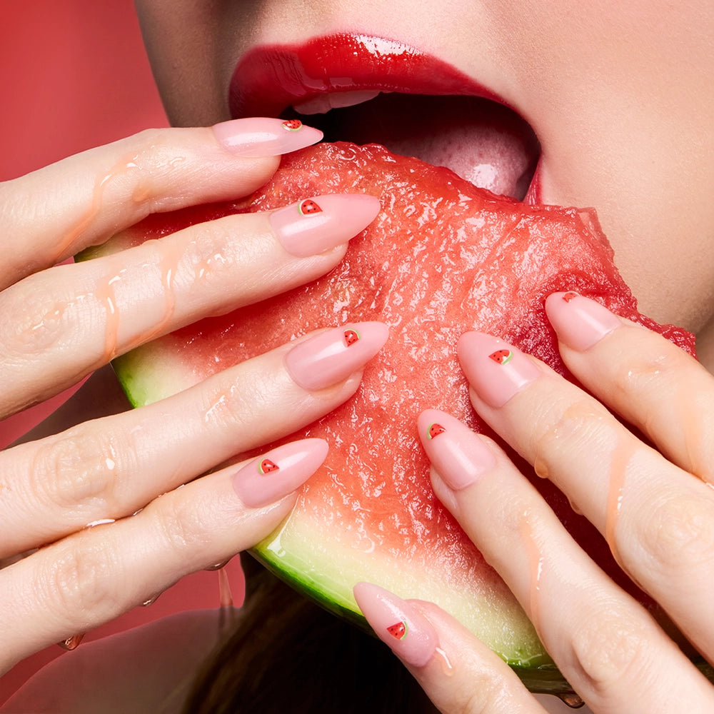 A woman with glossy red lips bites into juicy watermelon. Her almond nails, featuring watermelon-inspired Juicy Slice by glamnetic, are perfectly manicured for summer—effortlessly achieved using these press-on nails. - Product image