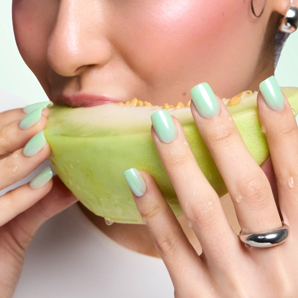 A person with glossy mint green Honeydewy nails by glamnetic and a silver ring holds and bites into a juicy melon slice. Only the lower part of their face and hands are visible. - Product image