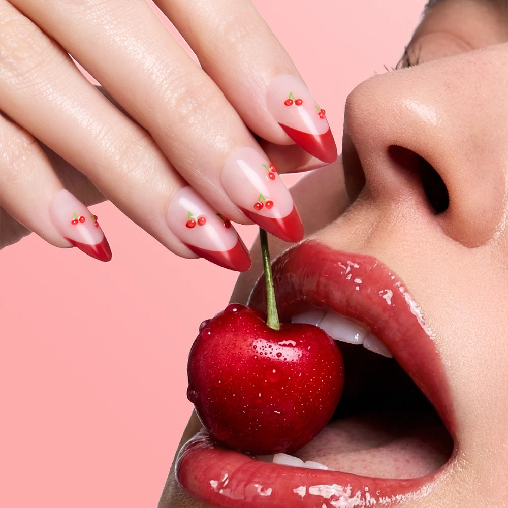 Glossy lips part near a red cherry held by manicured fingers with almond nails painted in glamnetic’s Cherry Bomb, featuring a cute cherry design, all set against a pink background. - Product image