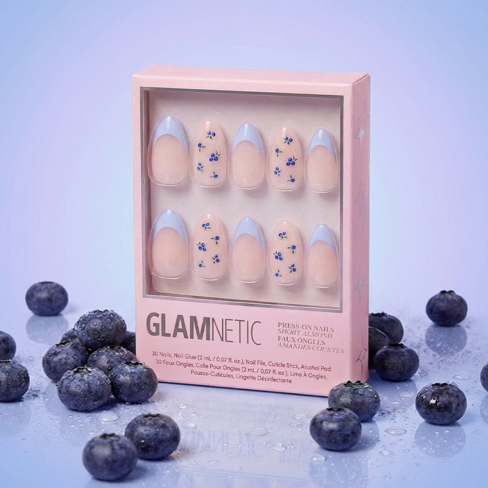 A box of glamnetic Bluebelle almond press-on nails, featuring pastel blue French tips and small blue floral designs, is displayed upright among fresh blueberries on a shiny light blue surface. - Product image