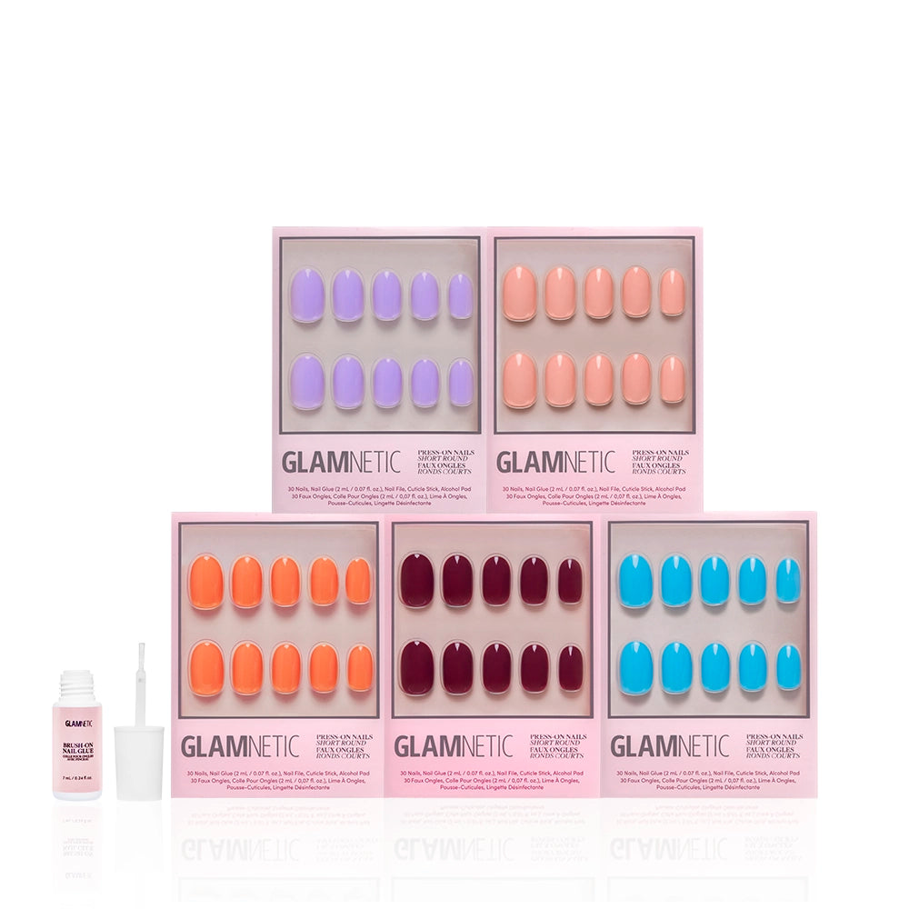 Five sets of glamnetic ‘Spring Round Up’ press-on nails in purple, coral, orange, and red are displayed in pink boxes. A small bottle of nail glue with an open cap sits on the left. Each set features short round glossy nails. - Product image