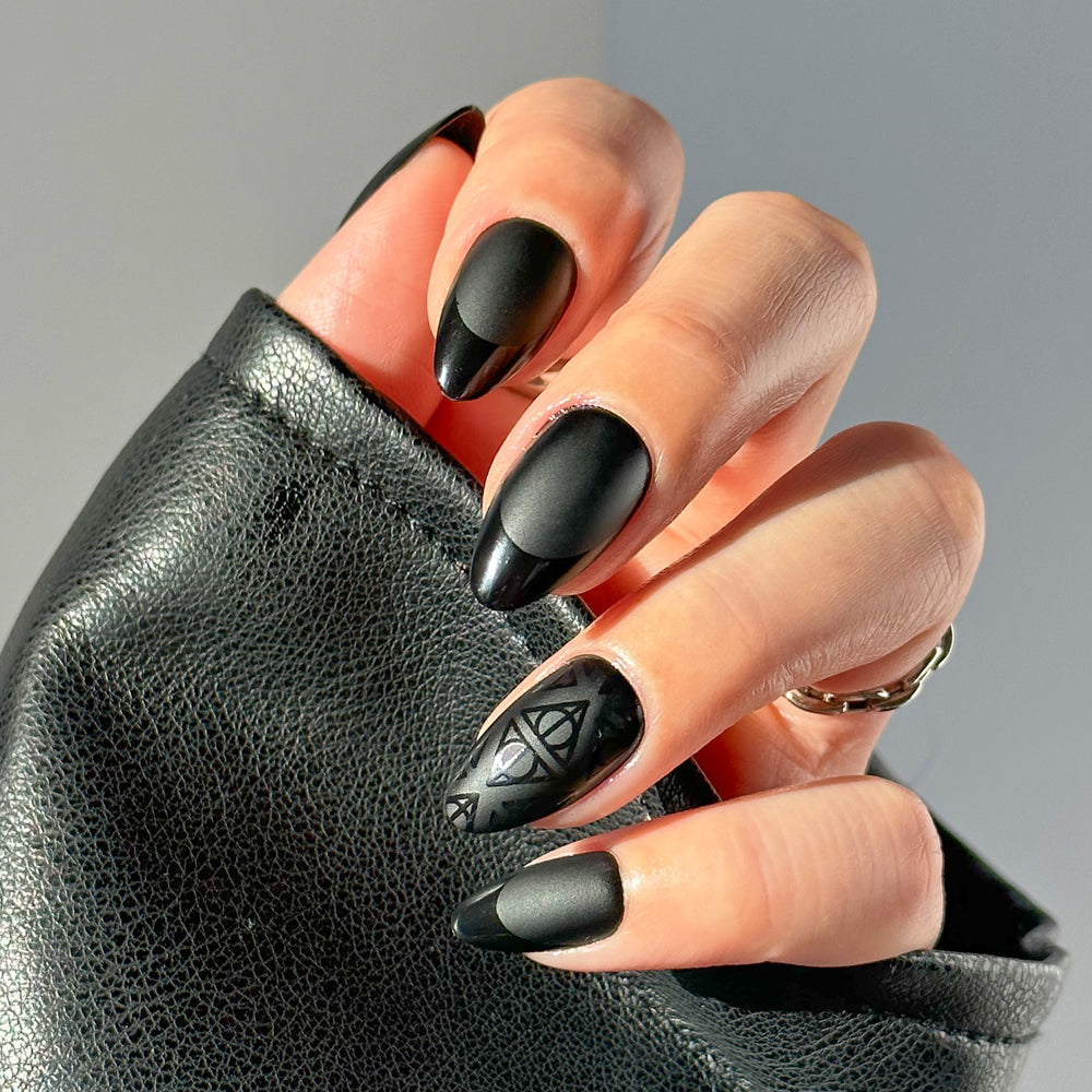A hand wearing glamnetic Deathly Hallows™ long, almond-shaped matte black press-ons—some with glossy tips and a geometric glossy accent—is partly covered by a black textured sleeve.