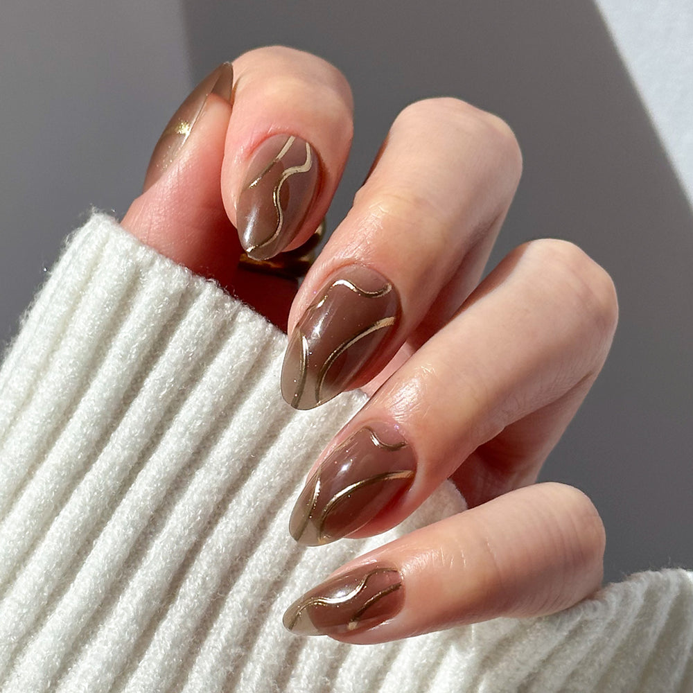 A hand with short almond nails, featuring glamnetic’s Golden Cup™ glossy brown polish and gold wavy lines, shown against a ribbed white sweater.