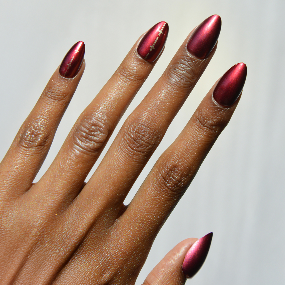 A hand with dark skin shows long, almond-shaped nails painted in glamnetic's Gryffindor™—a metallic burgundy polish. The ring and middle fingers feature subtle gold line designs, set against a soft gradient background.