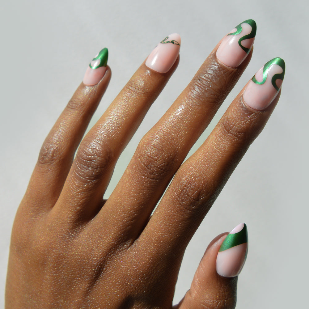 A hand with short almond nails showcases glamnetic's Parseltongue™ set—green and nude abstract designs, including curved green tips and swirling accents on a light nude base for a subtle Slytherin-inspired look.