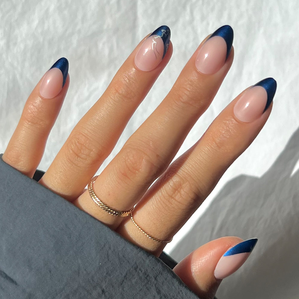 A hand with short almond-shaped nails showcases glamnetic's Ravenclaw™: a nude base with deep sapphire blue chrome French tips and a delicate white swirl accent. Thin gold rings adorn the fingers, partly covered by a gray sleeve.