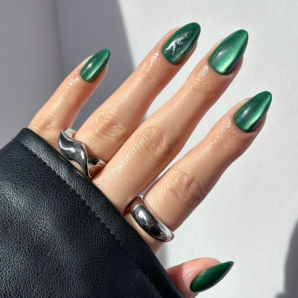 A hand with almond-shaped, shimmery emerald green glamnetic Slytherin™ nails with a subtle velvet finish and a white abstract accent design, plus two chunky silver rings and a black leather sleeve.