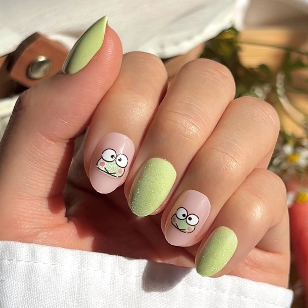 A hand with pastel green and nude nails shows off cute cartoon frog faces inspired by the glamnetic Keroppi™ Nail Set. Sunlight highlights this playful Sanrio design, and the person wears a white sleeve.