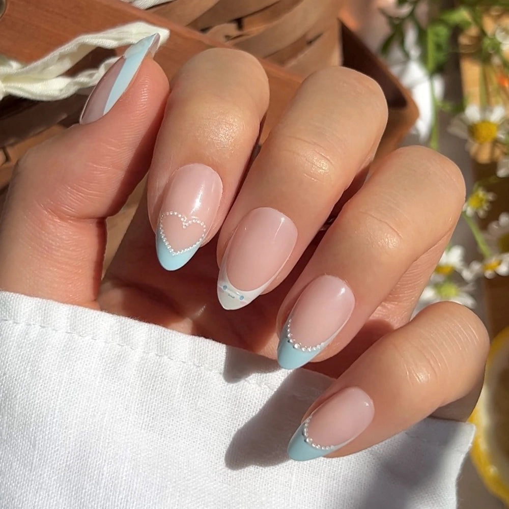 A hand with glamnetic's Cinnamoroll™ French Tip press-ons features light blue French tips, white beaded accents, and a pearl-outlined heart on the ring finger, all against a soft background and resting on a white sleeve.