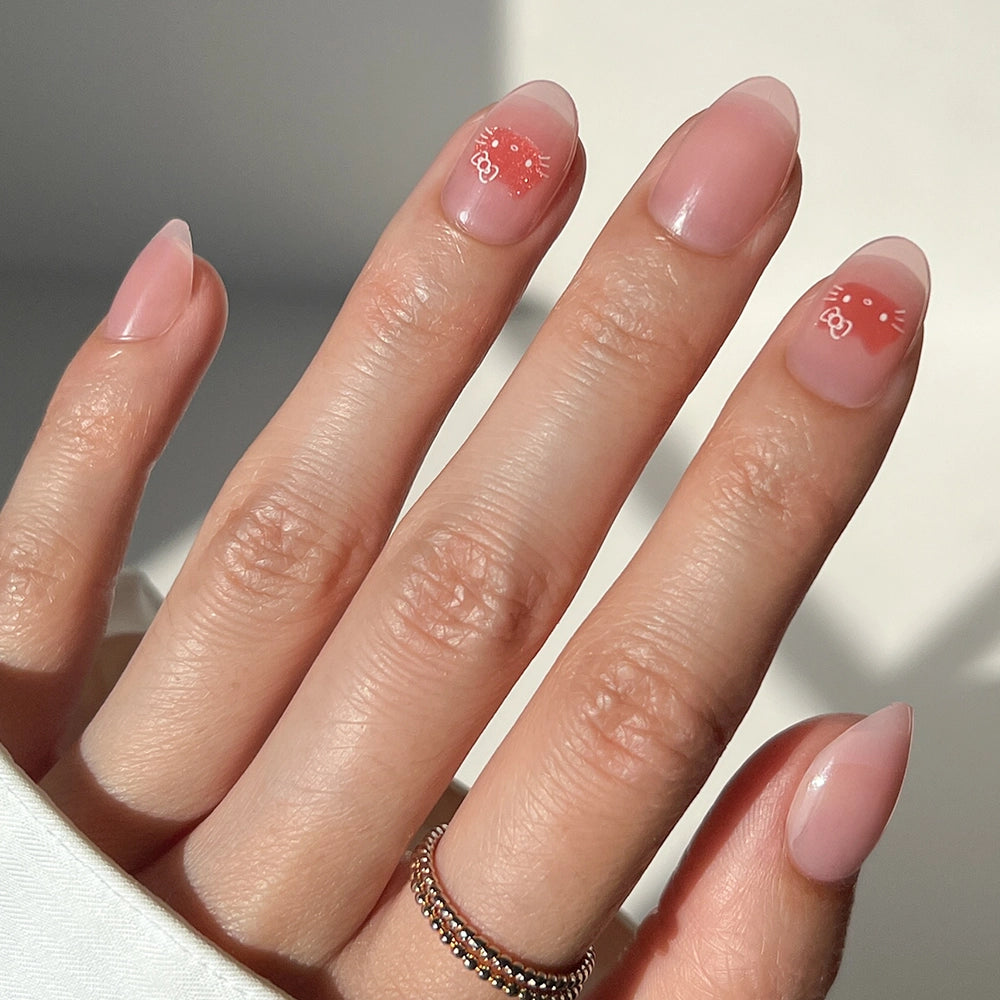 A hand with natural pink almond-shaped nails showcases two red Hello Kitty® designs by glamnetic, paired with a silver beaded ring on the middle finger.