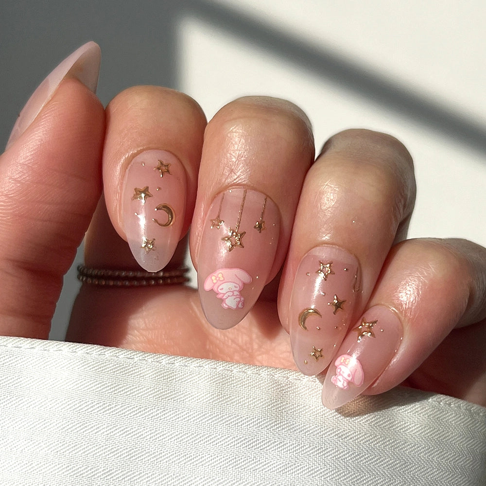 A hand with almond-shaped nails painted in sheer pink polish, adorned with gold star and crescent moon decals and small My Melody™ designs by glamnetic, rests on a light fabric.