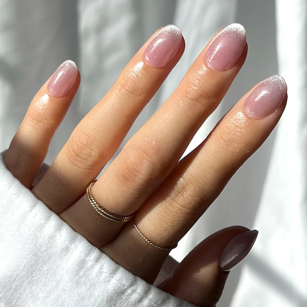 A hand with neatly manicured, almond-shaped nails in a soft pink base and frosty white tips from glamnetic’s Fairy Dust set, accented with two gold rings and a white long-sleeve shirt. - Product image