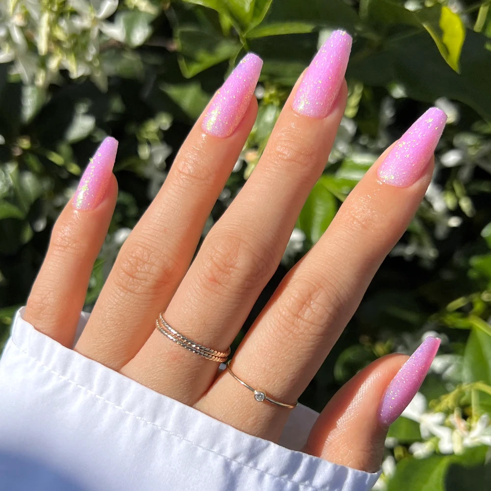 A hand with long, coffin-shaped pink nails wears “Juicy” press-ons by glamnetic, set against green leaves. A white sleeve and three gold rings highlight the glossy, elegant look of these eye-catching nails.