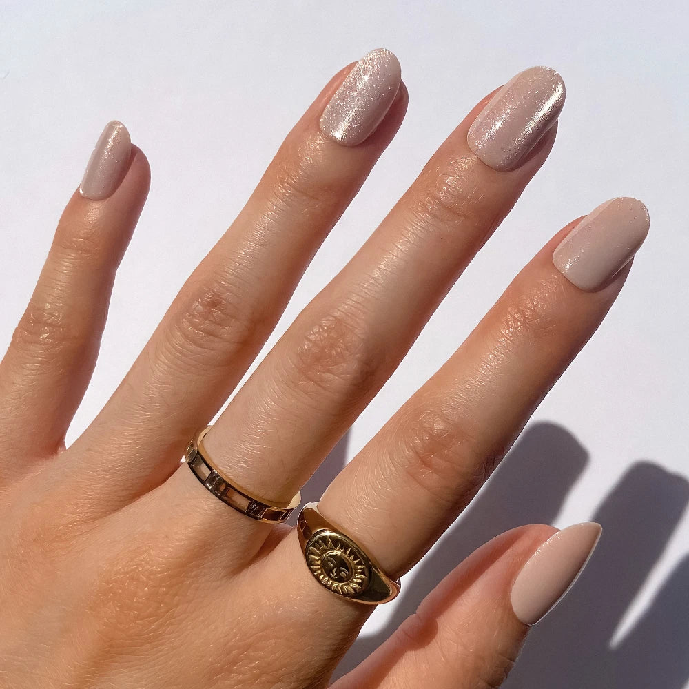 A hand with almond-shaped glamnetic Pure Intentions press-on nails in a pale nude shimmer shade, shown against a white background, is adorned with two gold rings—one plain and one featuring a sun design.