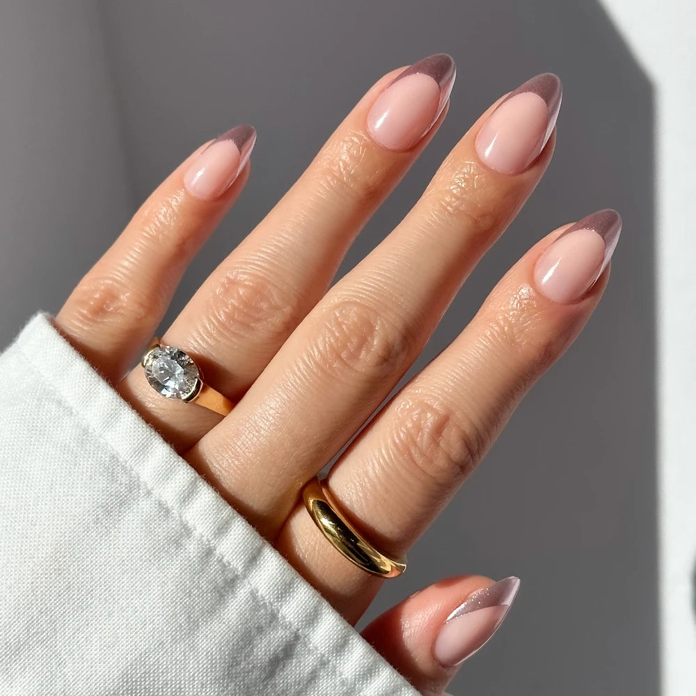 A hand with short almond nails wears glamnetic's Rock Candy, a pink and brown French manicure with chrome rose gold tips. The look is paired with a gold band, a large gemstone ring, and a white long-sleeve shirt.