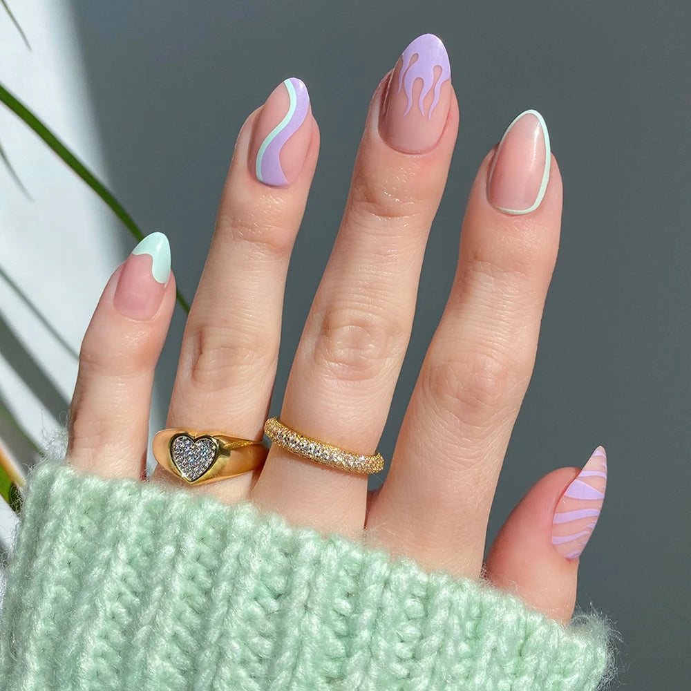 A hand with long, semi-transparent almond nails features glamnetic's Wild Card press-ons—pastel waves, flames, and stripes in mint green, lavender, and white—styled with gold rings and a mint knit sleeve. - Product image
