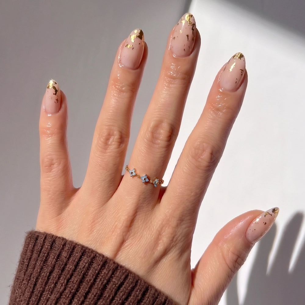 A hand with glossy nude short almond nails from glamnetic's 24K gold foil nail set is shown wearing a delicate gemstone ring and a brown ribbed sleeve, with a softly lit background adding an elegant touch. - Product image