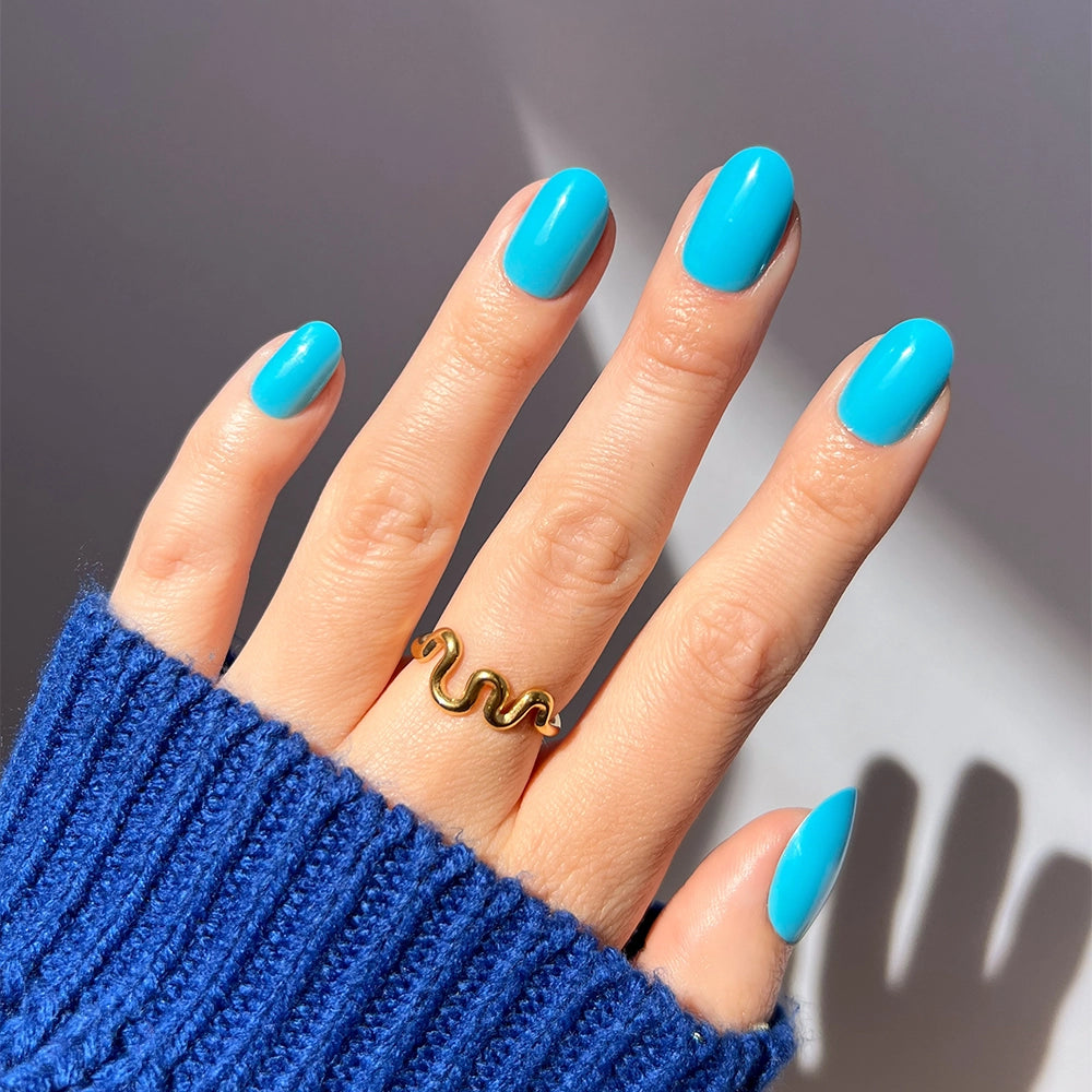 A hand wears glamnetic's Azure Blue short round press-on nails and a gold wavy ring on the middle finger. The royal blue sweater and sunlight in the background highlight the vibrant, long-lasting nail color.