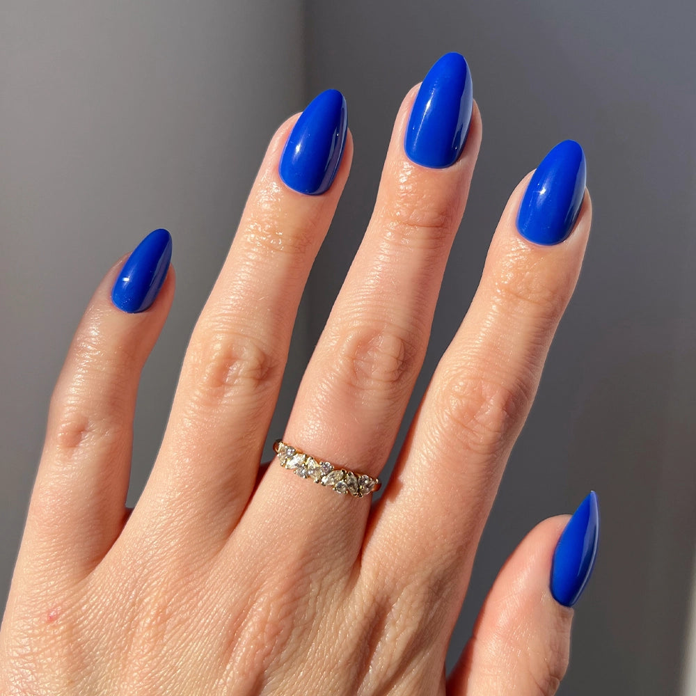 A hand with long, almond-shaped "Cobalt Blue" nails by glamnetic, wearing a gold ring with small diamonds on the ring finger, is displayed against a neutral background.