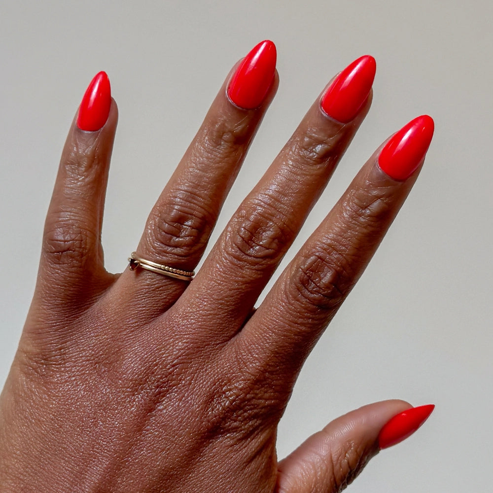 A hand with medium brown skin and short almond nails painted with glamnetic’s Fiery Red polish is shown. The ring finger has a thin, twisted gold ring. The background is plain and light-colored.