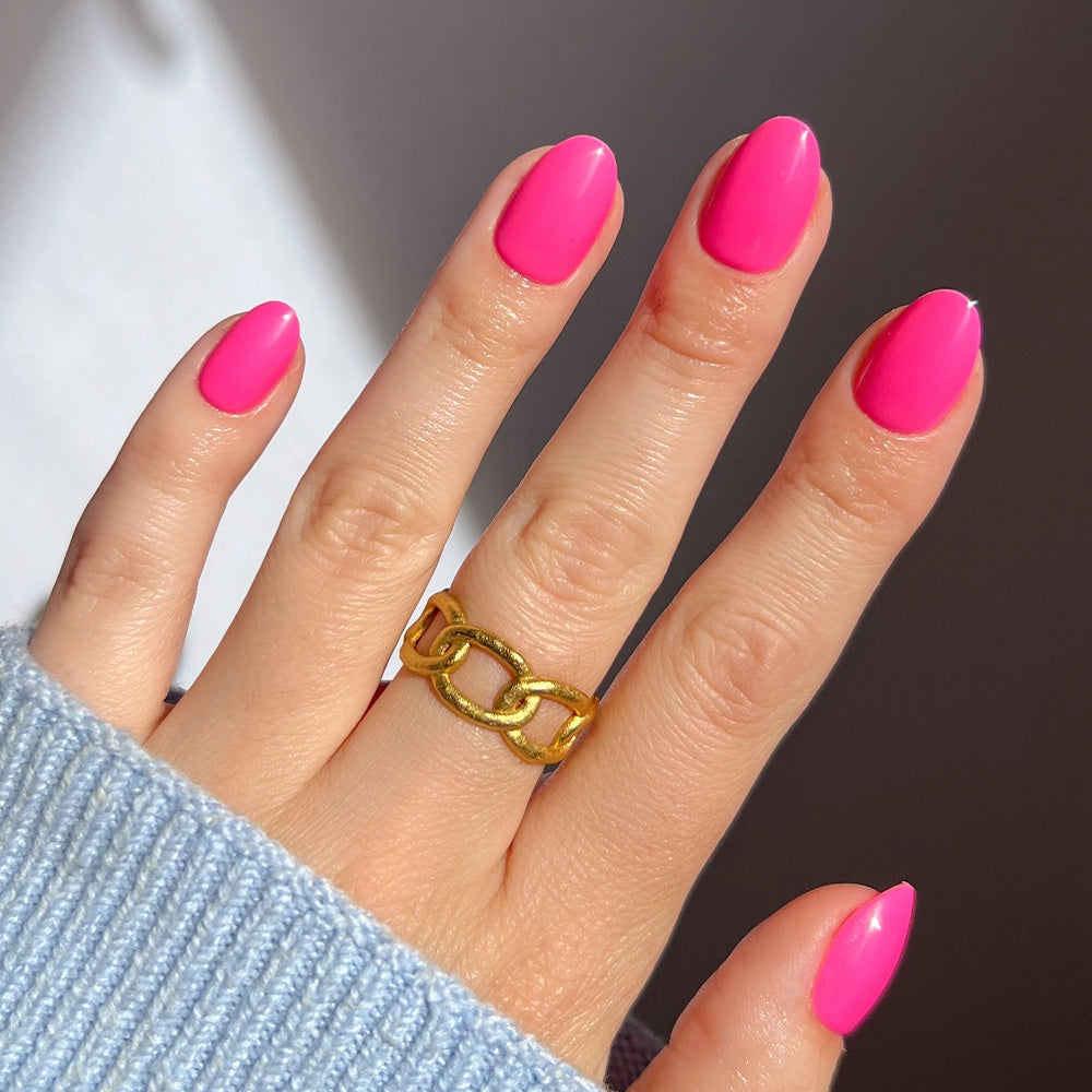 A hand with glossy, almond-shaped "Hot Pink" nails by glamnetic wears a chunky gold chain ring on the middle finger, paired with a light blue ribbed sweater. The softly lit background enhances the vibrant summer-ready look.