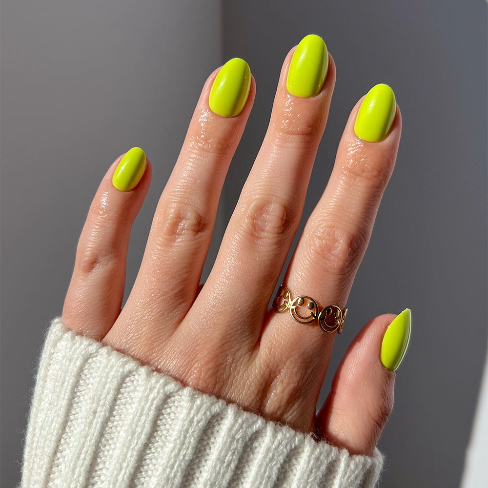 A hand with glamnetic Lime Green short oval press-on nails is shown, paired with a gold smiley ring and a white ribbed sweater. The softly lit background highlights the vivid color of these trendy nails. - Product image