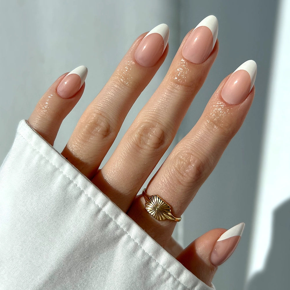 A hand with long almond-shaped Ma Damn nails by glamnetic features a glossy French manicure with white tips, paired elegantly with a gold sunburst ring and a white cuffed sleeve.