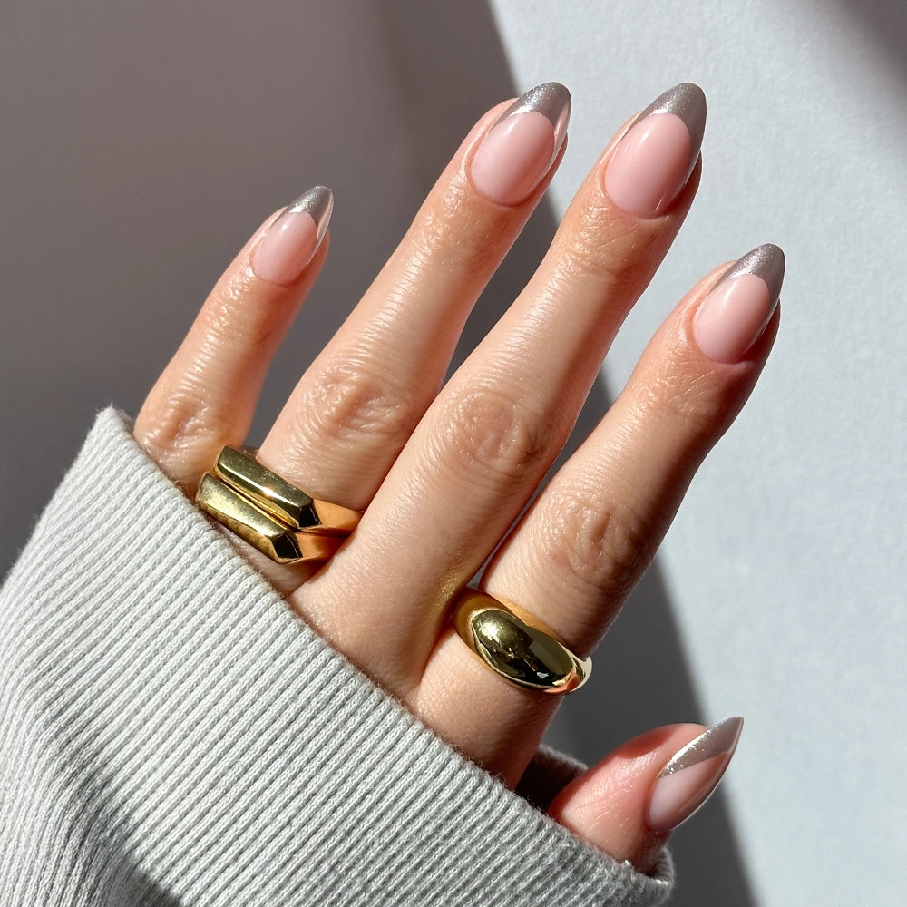A hand wears glamnetic's Mirror Mirror short almond press-on nails in light pink with chrome silver French tips, three chunky gold rings, and a light gray ribbed sleeve. - Product image