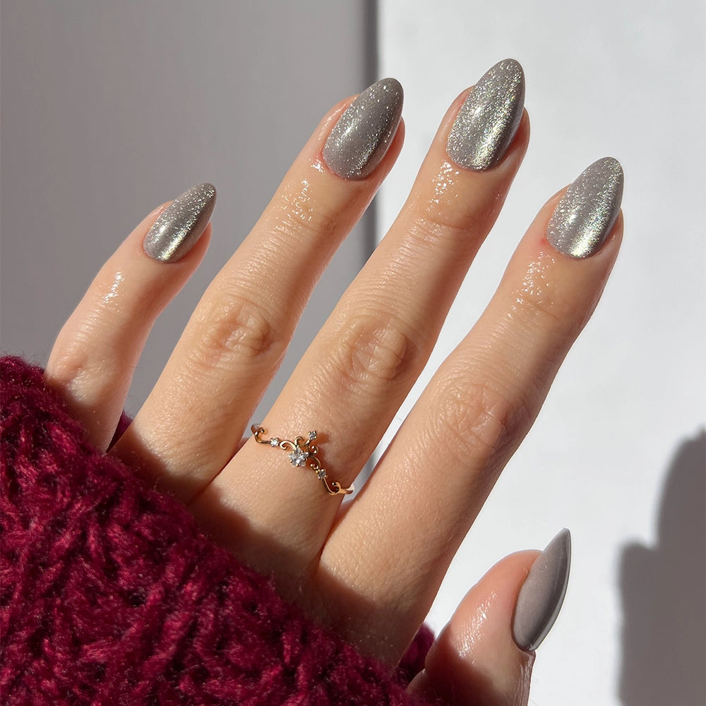 A hand with long, silver cat-eye glamnetic Oslo nails is shown, paired with a delicate gold ring and a chunky dark red knit sweater. The softly lit, neutral background highlights these vegan, cruelty-free nails for a stylish, conscious look. - Product image