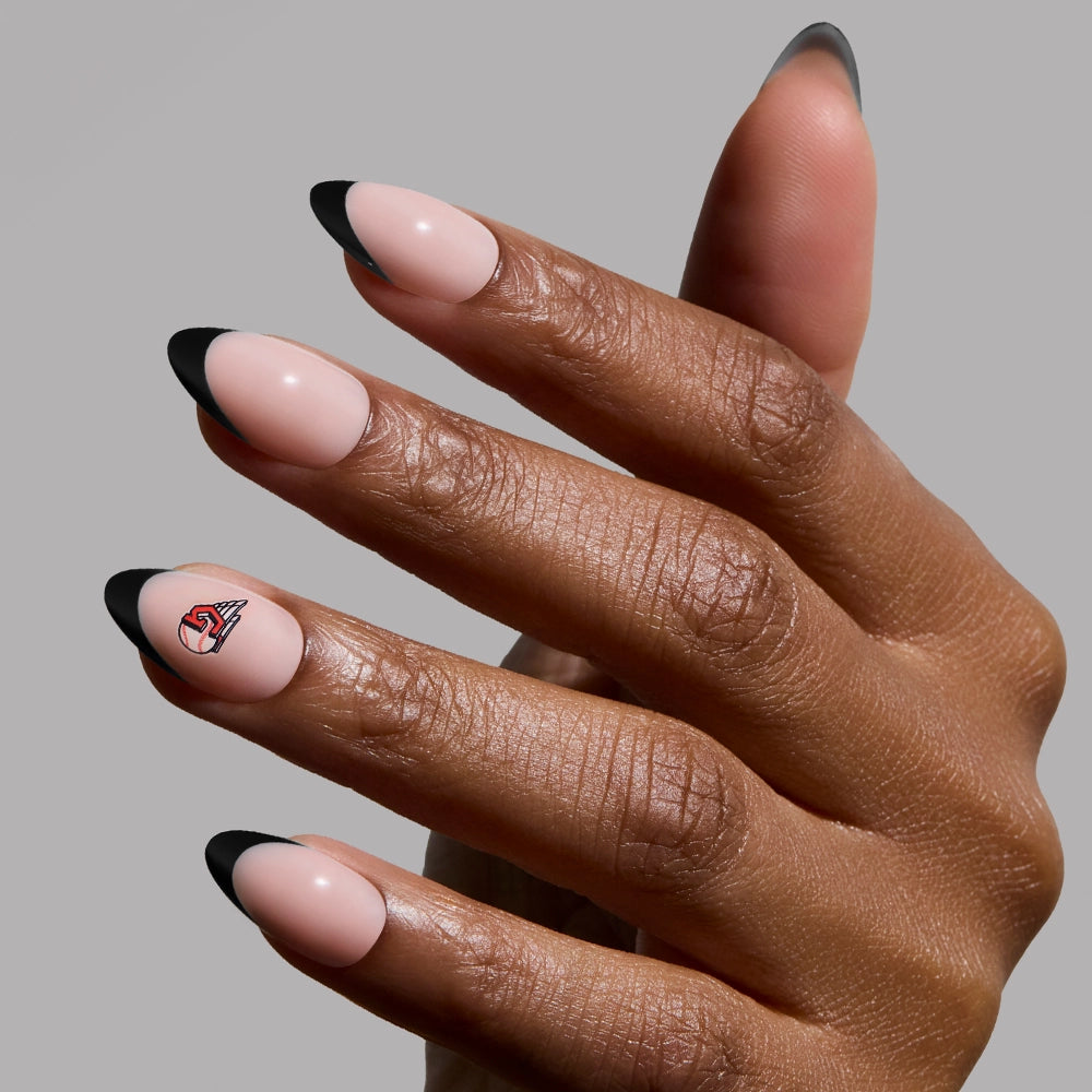 A hand with short almond nails painted nude with black French tips, plus a small red and black accent, resembles the glamnetic Cleveland Guardians Press-On Nail Set. The background is plain gray.