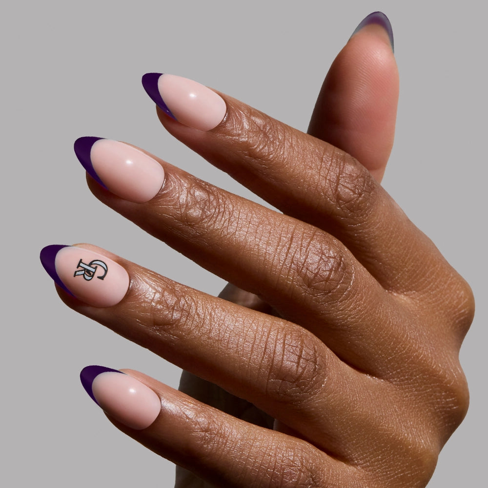 A hand with Colorado Rockies Press-On Nail Set by glamnetic shows long almond nails with a nude base, dark purple French tips, and a black-and-white graphic on the ring finger, set against a light gray background.
