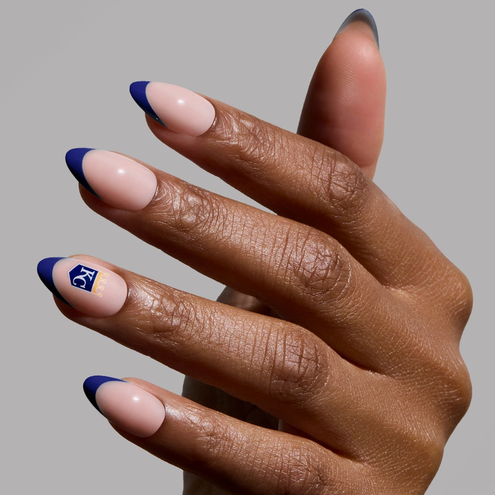 A hand with almond-shaped nude nails and dark blue tips, plus a small blue square with white text on the ring finger, showcases the glamnetic Kansas City Royals Press-On Nail Set against a plain gray background.