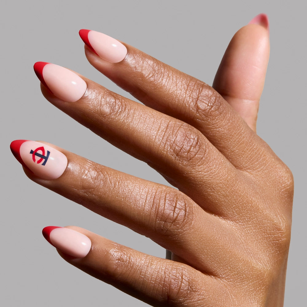 A hand with glossy almond nails featuring a nude base and red French tips; the ring finger displays the glamnetic Minnesota Twins Press-On Nail Set in red and navy blue. The background is plain gray.
