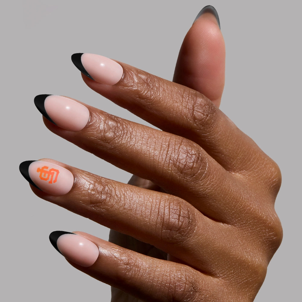 Show off your team spirit with the glamnetic San Francisco Giants Press-On Nail Set, featuring almond-shaped nude nails with black French tips and an orange SF logo—perfect for fans, shown against a plain gray background.