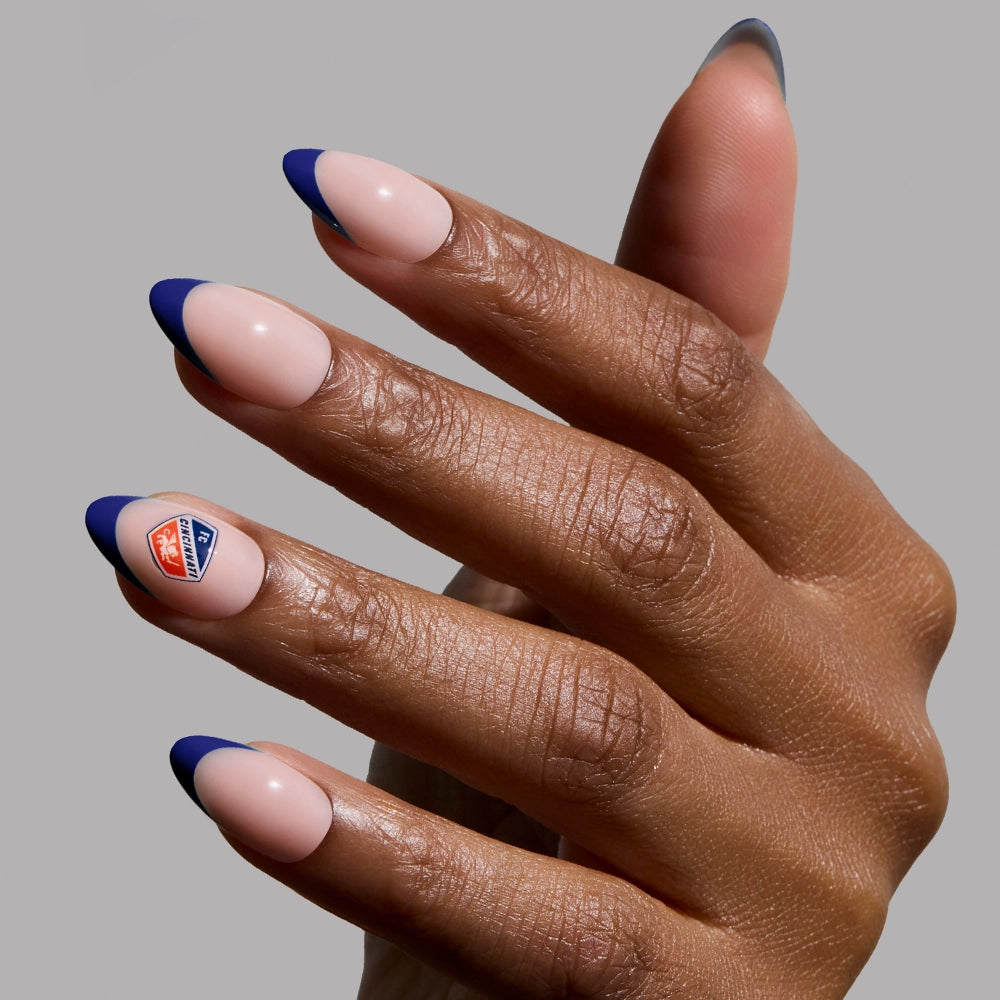 A hand with almond vegan press-on nails from glamnetic’s FC Cincinnati Press-On Nail Set features a nude base, dark blue tips, and an "I Voted" sticker on the ring finger. The plain gray background highlights their chic look.