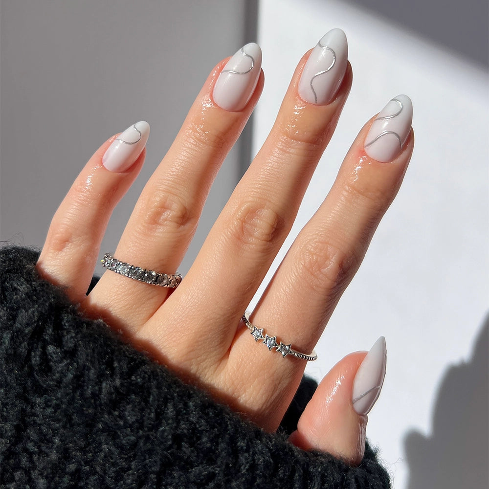 A hand with short almond nails in glamnetic’s Silent Night—a glossy pale pink polish with white abstract lines. Three silver rings and a black textured sweater highlight these trendy vegan nails. - Product image
