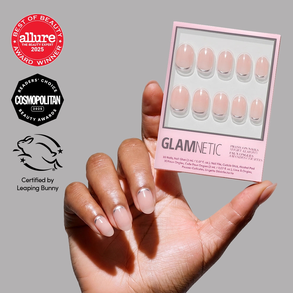 A hand with neatly manicured short round nails holds a pink box of glamnetic Silver Belle press-on nails. The box displays various nail sizes and features award badges from Allure, Cosmopolitan, and Leaping Bunny certification on the side.
