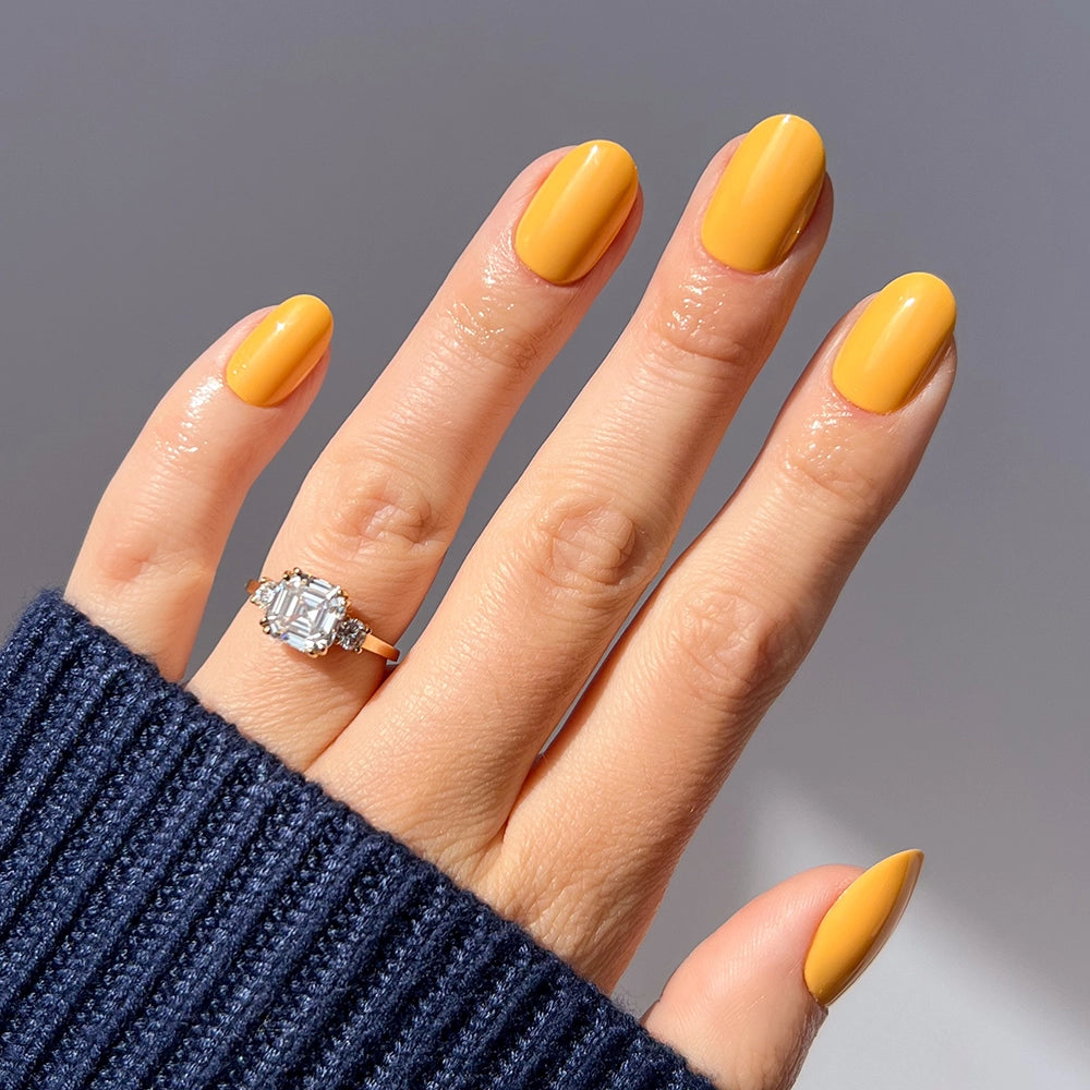 A hand with neatly manicured glamnetic Sunset Orange press-on nails wears a large, rectangular-cut diamond ring on the ring finger, partially covered by a navy blue ribbed sweater.