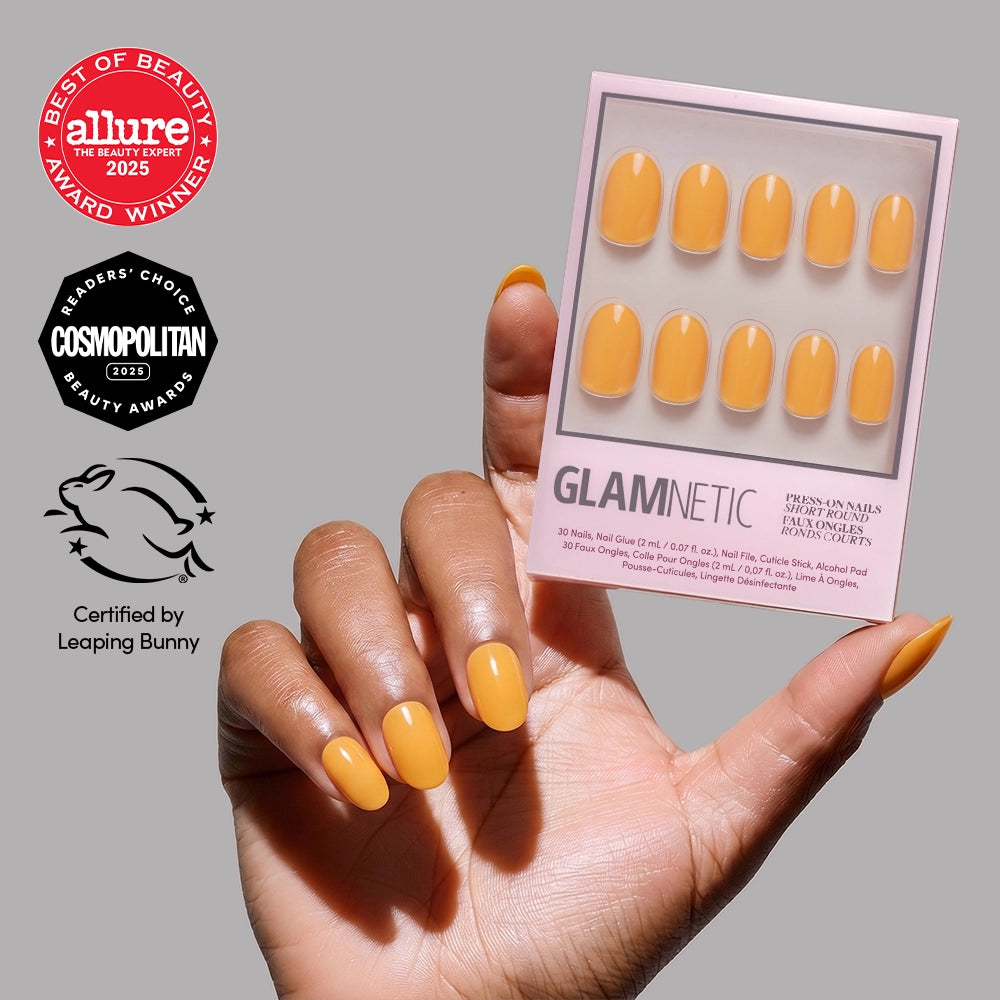 A hand with neatly manicured short round nails in Sunset Orange holds a box of glamnetic press-on nails in the matching shade. Allure, Cosmopolitan, and Leaping Bunny certification badges are displayed on the left.