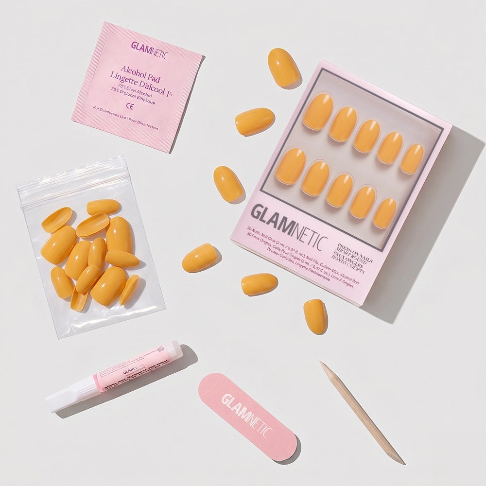 A set of Sunset Orange short round press-on nails with glue, cuticle stick, nail file, alcohol pad, and extra nails are arranged on a white surface. The packaging and tools feature glamnetic branding—a perfect summer nail color. - Product image