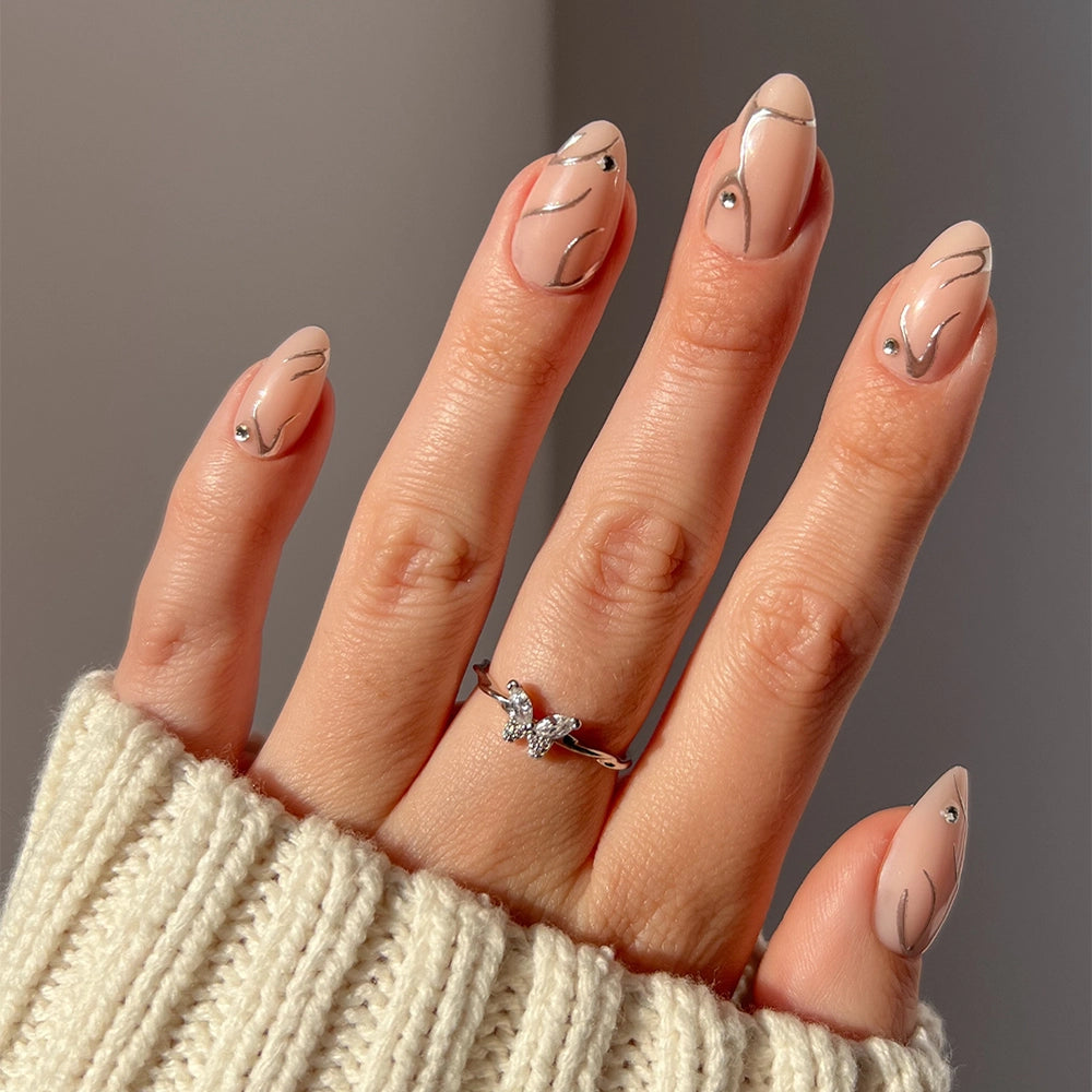 A hand with short almond nude nails features minimal black lines and small rhinestones. Styled with a silver butterfly ring, a cream knit sweater, and flaunting the glamnetic Astro Nail Set.