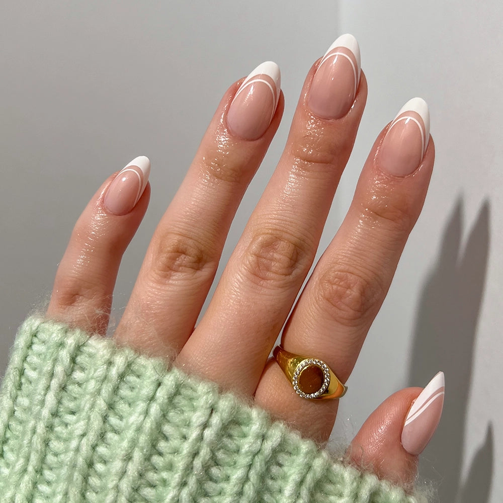 A hand with short almond nails wears glamnetic's Paris press-ons, showcasing a modern French tip with white accents. The look is styled with a gold ring set with a dark stone and finished off by a pale green knit sweater. - Product image