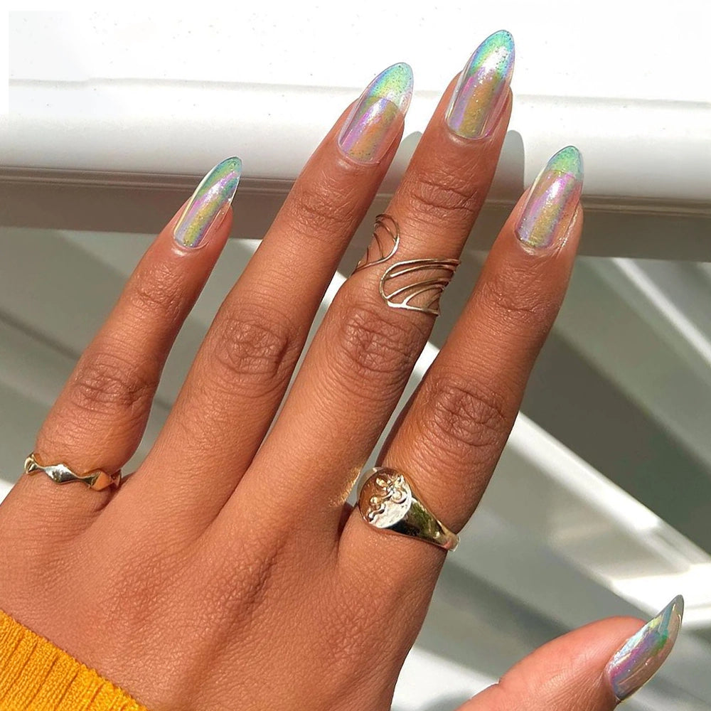 A hand wearing three gold rings shows off glamnetic’s Stardust iridescent holographic almond press-ons against a blurred white background, paired with a yellow long-sleeve top.